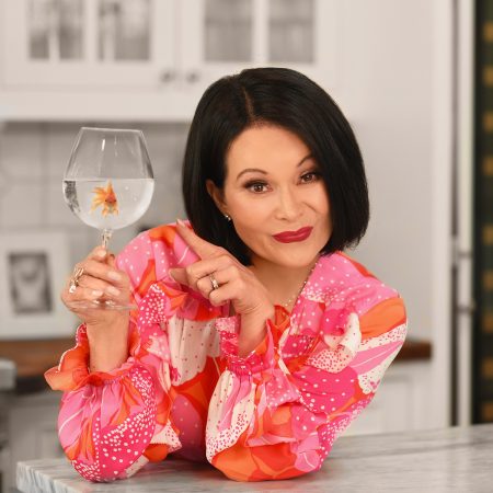 Tracy is in her kitchen, holding up a wine glass filled with water & a goldfish & pointing to it